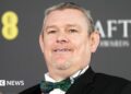 Kirk Jones in a tuxedo at the Baftas, he has glasses and grey, wavy hair