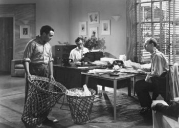 Black-and-white image of two men sitting at a table and a man emptying a bin