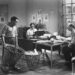 Black-and-white image of two men sitting at a table and a man emptying a bin