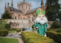 An individual dressed in a green ecclesiastical robe with a white beard and green mitre stands in front of a historical building with a Gothic architectural style, positioned amidst a meticulously maintained garden.