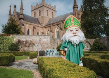 An individual dressed in a green ecclesiastical robe with a white beard and green mitre stands in front of a historical building with a Gothic architectural style, positioned amidst a meticulously maintained garden.