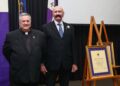 Two individuals in formal attire stand beside an easel displaying a framed certificate.