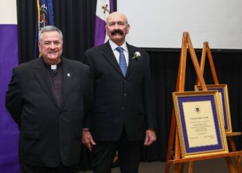 Two individuals in formal attire stand beside an easel displaying a framed certificate.