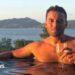 Jordan Wright holding a wine glass at the edge of an infinity pool. He has brown hair and stubble and the sun is shining on him. Tall green hills and the sea are pictured behind him.