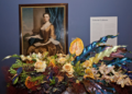 The image shows a framed portrait of a woman in historical attire, displayed on a blue wall with a floral arrangement of roses and glossy leaves beneath it.
