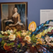 The image shows a framed portrait of a woman in historical attire, displayed on a blue wall with a floral arrangement of roses and glossy leaves beneath it.