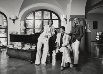 Three individuals in stylish attire pose in a sophisticated interior with a piano.