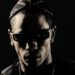 Travis Scott with braided hair and sunglasses, set against a dark background.