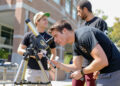 Man wearing black shirt bends down to look through telescope. There are two other men standing behind him talking.