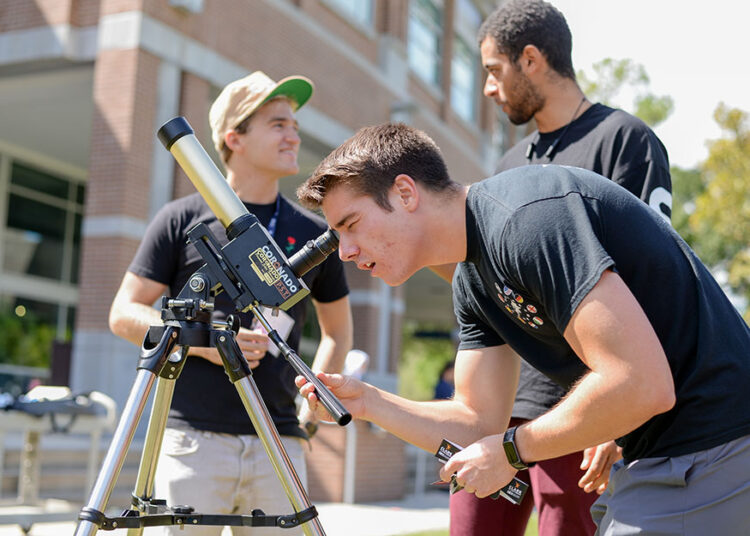 Man wearing black shirt bends down to look through telescope. There are two other men standing behind him talking.