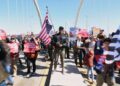 Kymone Freeman holds an American flag while demonstrators stream past him over the Frederick Douglass Bridge during the No Kings protest in Washington, Saturday, March 28, 2026. (AP Photo/Tom Brenner)