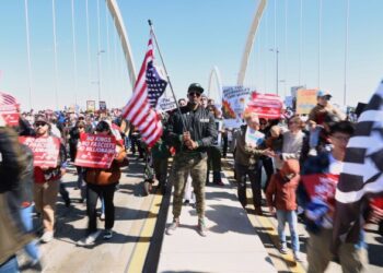 Kymone Freeman holds an American flag while demonstrators stream past him over the Frederick Douglass Bridge during the No Kings protest in Washington, Saturday, March 28, 2026. (AP Photo/Tom Brenner)