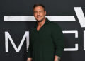 Alan Ritchson, in a dark green sweater, poses confidently against a dark backdrop with large white letters.