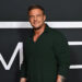 Alan Ritchson, in a dark green sweater, poses confidently against a dark backdrop with large white letters.