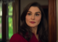 A woman in a red blouse stands in a warmly lit indoor setting.
