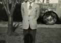 A young boy in a suit stands on grass with his hands behind his back, in front of a tree and a car, with houses visible in the background. The photo is black and white.