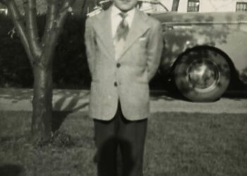 A young boy in a suit stands on grass with his hands behind his back, in front of a tree and a car, with houses visible in the background. The photo is black and white.