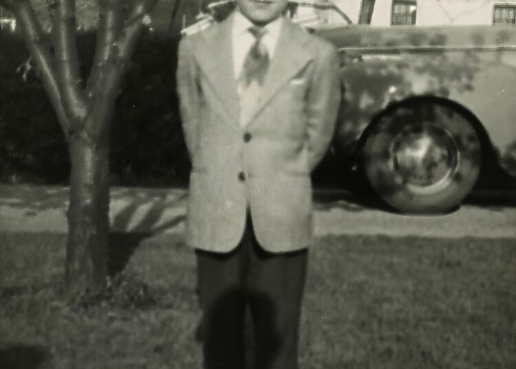 A young boy in a suit stands on grass with his hands behind his back, in front of a tree and a car, with houses visible in the background. The photo is black and white.