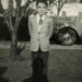 A young boy in a suit stands on grass with his hands behind his back, in front of a tree and a car, with houses visible in the background. The photo is black and white.