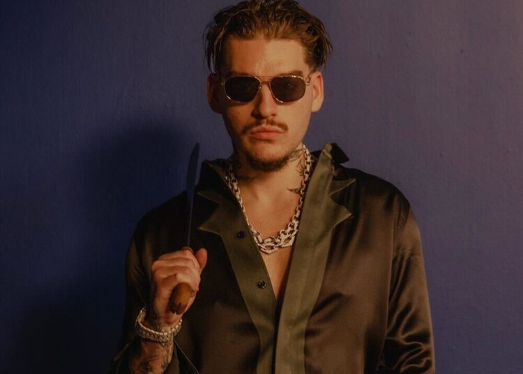 Rapper Yeat wearing sunglasses and a dark satin shirt holding a knife, posing against a blue background. This is a photo attached to his latest album, ADL, which is a double album.