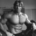 Shirtless Arnold Schwarzenegger with one hand on his hip and the other resting on a barbell