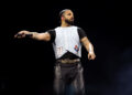 Drake performing on stage, wearing a white vest and black shirt, holding a microphone. He is getting ready to release 'Iceman' his latest album.