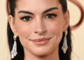 Anne Hathaway smiling, wearing elegant earrings, with her hair pulled back, in a close-up portrait.