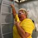 An individual in a yellow shirt and white goggles is leaning against a ladder affixed to a wall, possibly engaged in a maintenance task.