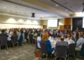 Large conference banquet with attendees seated at round tables facing a speaker and projection screens.