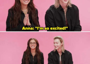 Two people sit against a pink background, smiling. One has long hair and glasses, wearing a dark jacket. The other has shoulder-length hair, also in a dark jacket