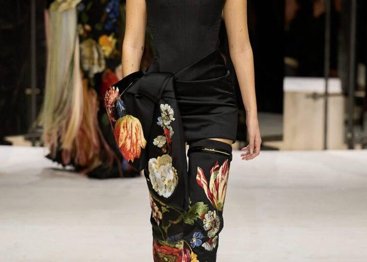 Fashion model wearing an elegant outfit with floral designs, walking on a runway.