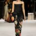 Fashion model wearing an elegant outfit with floral designs, walking on a runway.