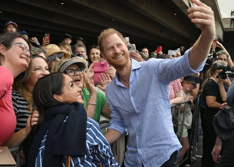 Prince Harry smiling and taking a selfie with a crowd of fans while Meghan Markle stands beside him during a public engagement.