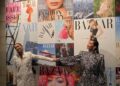 Two individuals posing in front of a collage of magazine covers.