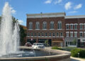A circular fountain in front of a brown brick building in Bryan, Ohio, with cars and trees in the background under a blue sky