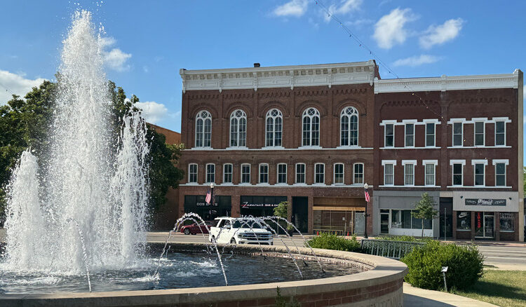 A circular fountain in front of a brown brick building in Bryan, Ohio, with cars and trees in the background under a blue sky
