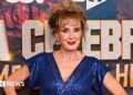 Beverley Callard in a blue dress with her hands on her hips, standing in front of a sign for I'm a Celebrity... South Africa