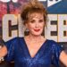 Beverley Callard in a blue dress with her hands on her hips, standing in front of a sign for I'm a Celebrity... South Africa