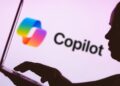 In this photo illustration, the Microsoft Copilot logo is seen in the background next to a silhouette of a person using a notebook.