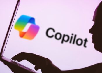 In this photo illustration, the Microsoft Copilot logo is seen in the background next to a silhouette of a person using a notebook.