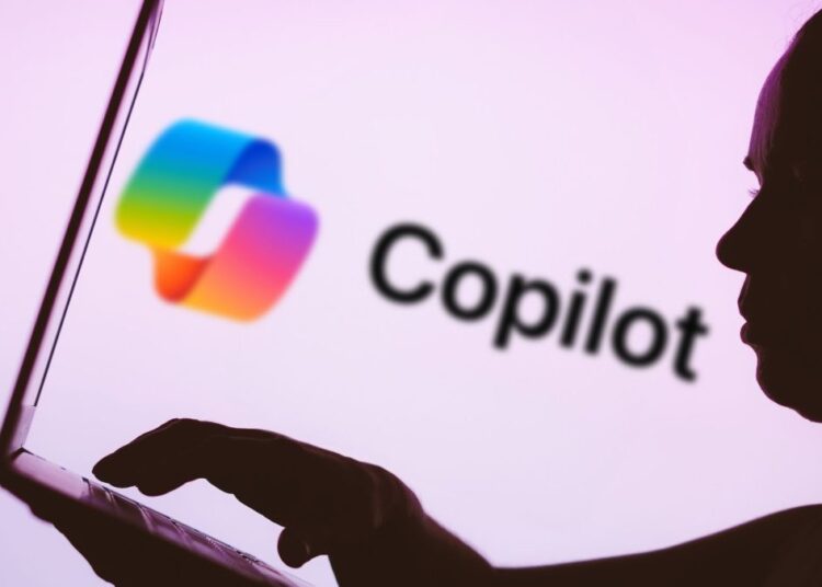 In this photo illustration, the Microsoft Copilot logo is seen in the background next to a silhouette of a person using a notebook.