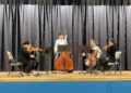 Five students are assembled on a stage. Two hold cellos – one is standing – while the other musicians, seated, play violin.