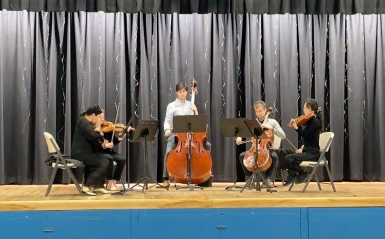 Five students are assembled on a stage. Two hold cellos – one is standing – while the other musicians, seated, play violin.