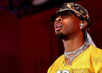 D4vd wearing a yellow sports jersey, sunglasses, and a patterned headscarf, with earrings and layered necklaces, looking upwards.