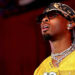 D4vd wearing a yellow sports jersey, sunglasses, and a patterned headscarf, with earrings and layered necklaces, looking upwards.