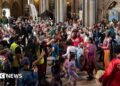 A large group of people dancing inside a church hall to a band. There is bunting across some of the internal archways in the church and there are lots of children among the dancers