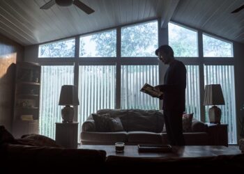 A person reading a magazine in a living room with large windows.