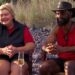 Two individuals are seated on chairs outdoors, positioned near a natural backdrop that includes a mix of rocks and vegetation. The individual on the left wears a red shirt and a dark vest, while the individual on the right is dressed in a red sleeveless shirt and a wide-brimmed hat. They are engaged in conversation and appear to be in a relaxed setting.