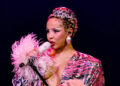 A performer singing on stage, wearing a sparkly outfit with feathered sleeves and a leopard-print headpiece, holding a microphone.