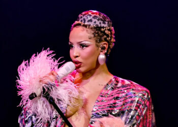 A performer singing on stage, wearing a sparkly outfit with feathered sleeves and a leopard-print headpiece, holding a microphone.